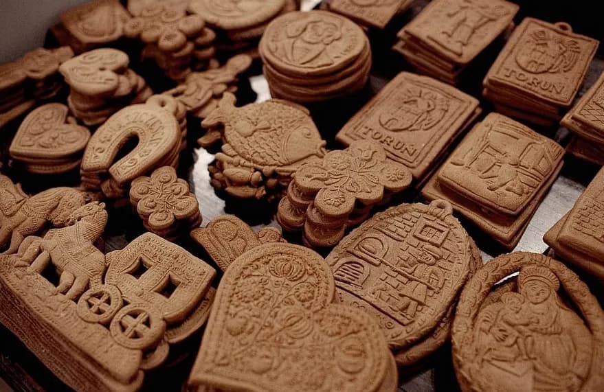 A close-up view of numerous intricately embossed gingerbread cookies in various shapes, including hearts, horses, fish, and historical scenes, stacked and scattered on a dark surface.