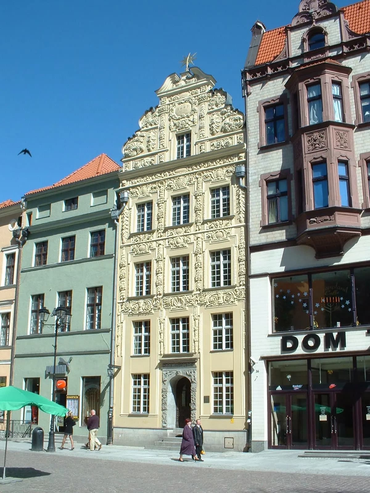 A tall, ornate yellow building with many windows stands prominently on a street, flanked by a simpler green building on the left and a building with a bay window and a storefront on the right.