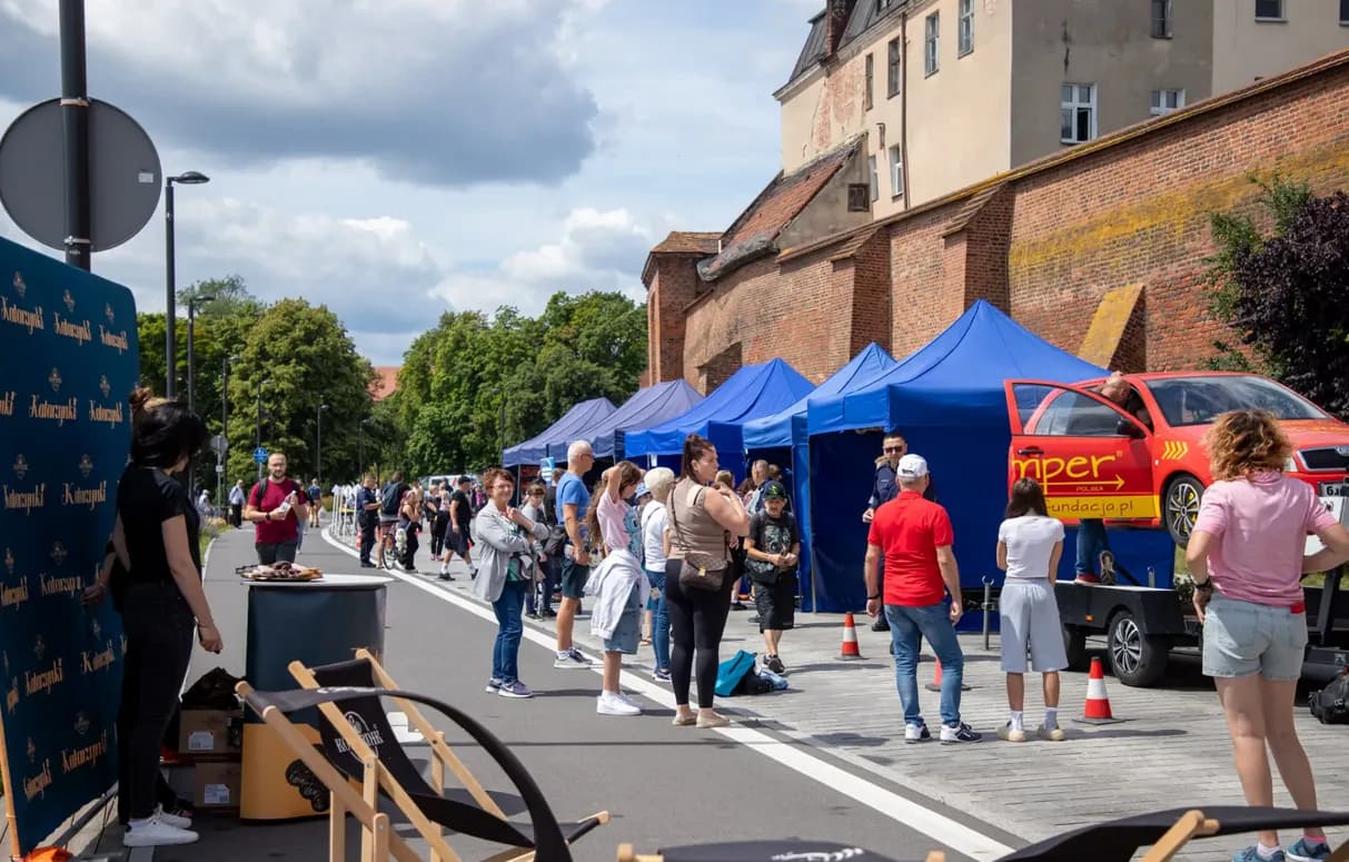 A street fair is in progress with many people gathered around several blue tents and an orange car on display, all set against a backdrop of historic buildings and green trees under a cloudy sky.