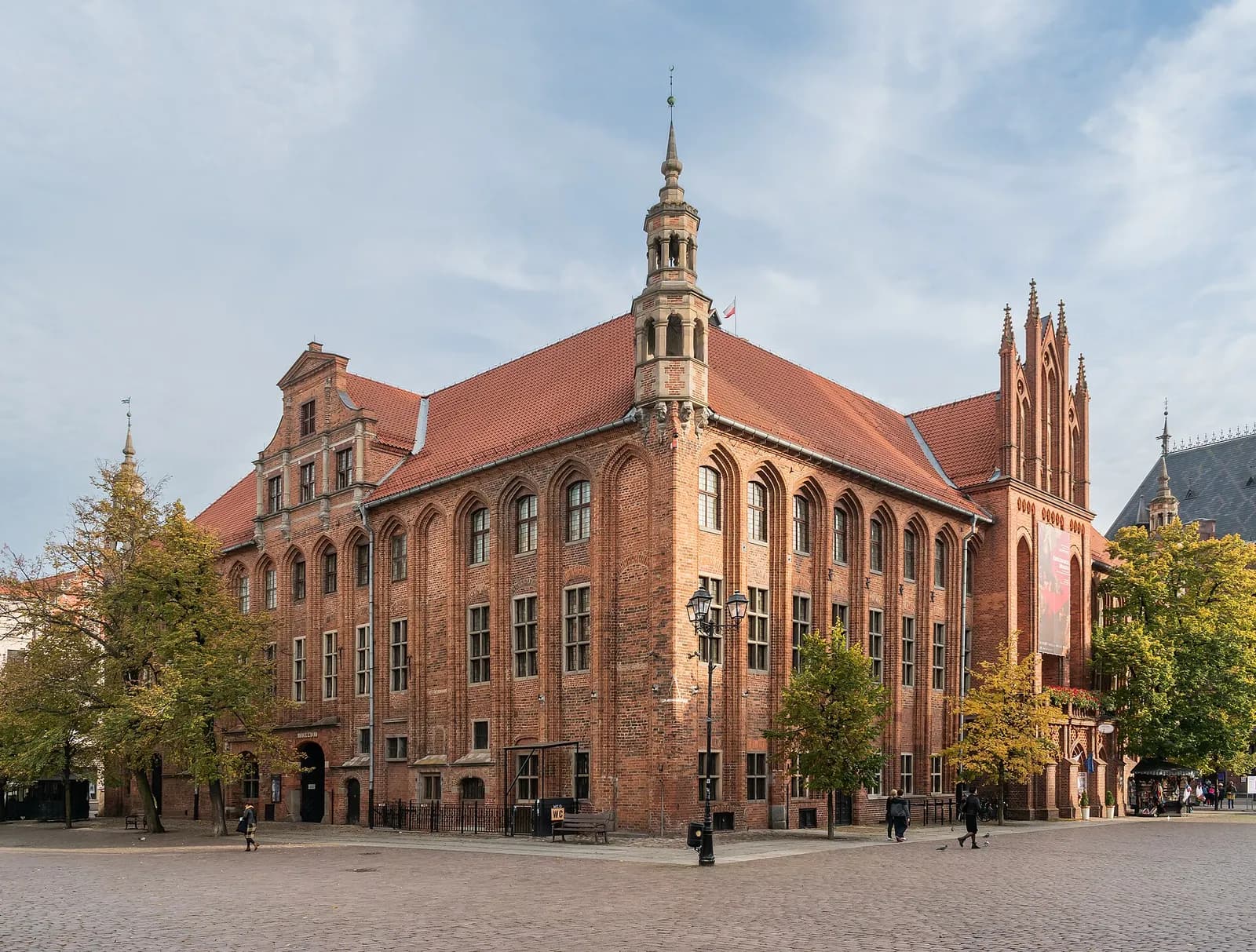 A large, multi-story brick building with arched windows and a prominent central spire stands in a cobblestone square under a partly cloudy sky.
